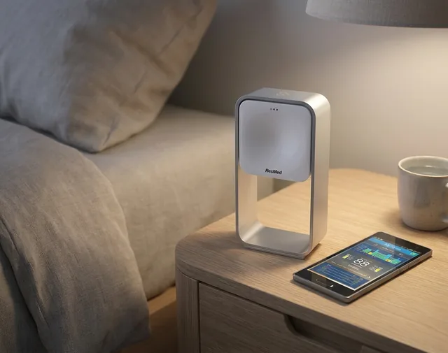 The ResMed S+ connected sleep device on a bedside table at night, its companion app open on a phone beside it.