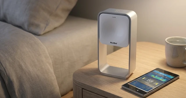 The ResMed S+ sensor on a bedside table beside a bed, with a phone next to it showing a night's sleep data.